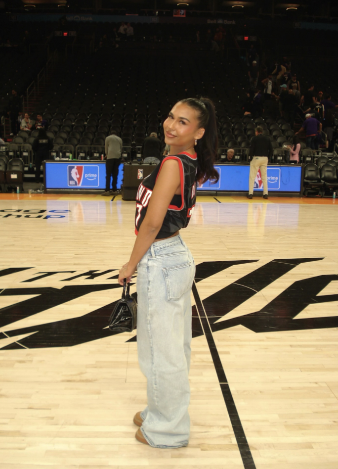 Woman on a basketball court wearing a fitted red jersey with high-waisted light-wash jeans and a small black bag.