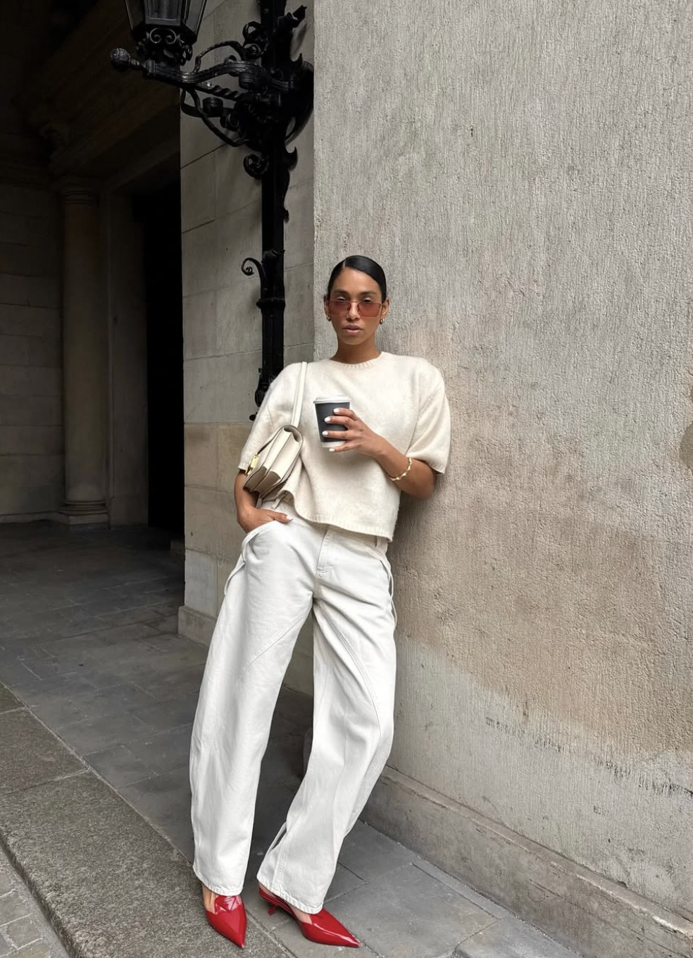 A woman leaning against a stone pillar wearing white straight-leg cargo jeans, red pointed-toe heels, and a cream oversized sweater while holding a coffee.