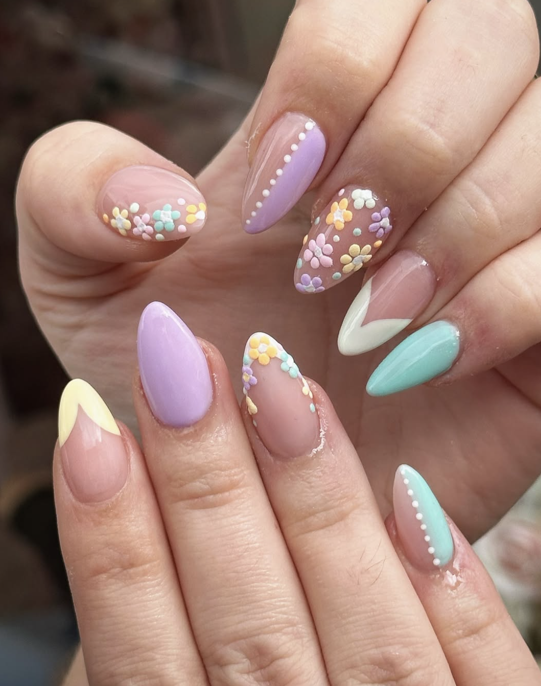 : A close-up of long stiletto-shaped acrylic nails featuring a variety of spring designs, including 3D textured flowers, yellow and white double-V French tips, and white polka dot "cuffs" on pastel lavender and mint green bases.