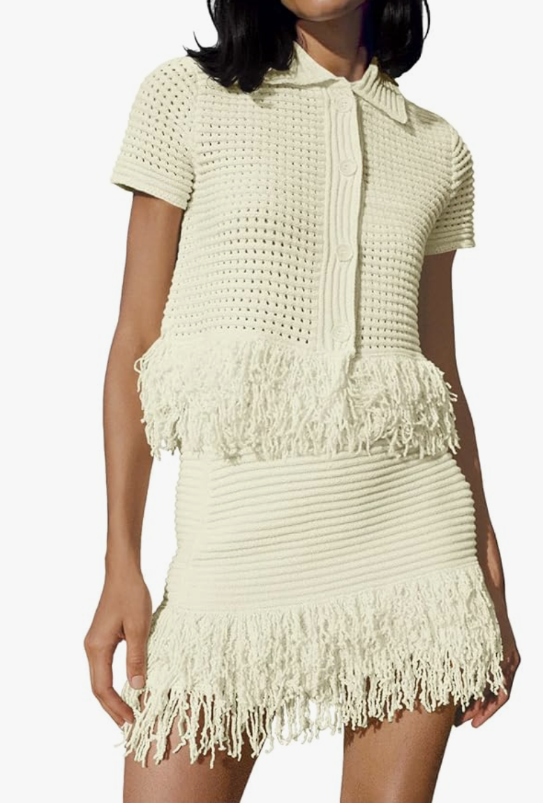 A woman wearing a cream-colored crochet two-piece set featuring a short-sleeve button-up top and a matching mini skirt with a long fringe hem.