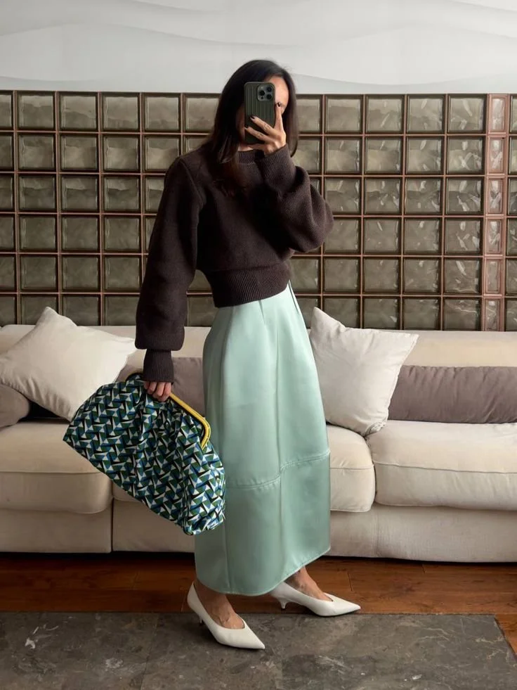 A fashion editor-approved look for Spring 2026, featuring a structured Jade green midi skirt paired with a cropped dark chocolate brown sweater, white pointed-toe heels, and a patterned clutch.