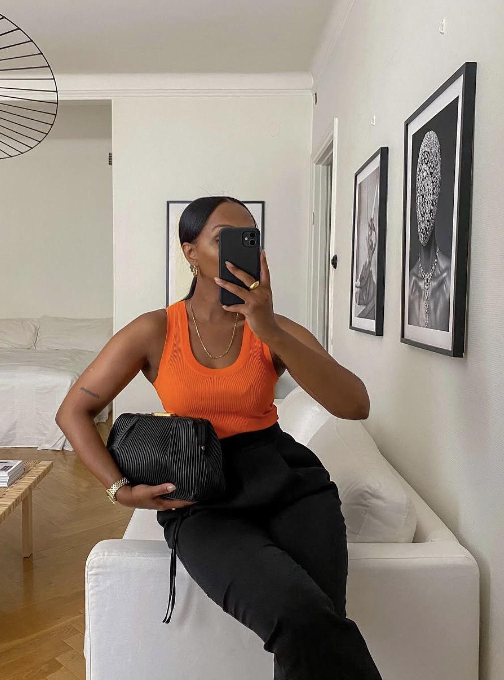 A woman wearing a vibrant orange ribbed tank top tucked into high-waisted black trousers, holding a black pleated clutch bag.