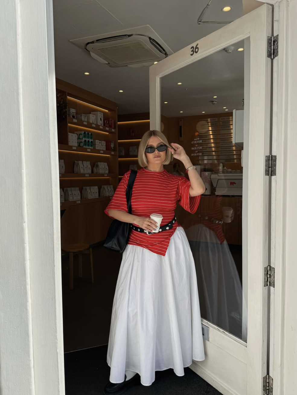 A woman posing in a white maxi skirt, a red and white striped t-shirt, and a black grommet belt, holding a coffee cup.