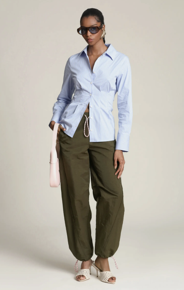 A woman wearing a blue and white striped button-down shirt styled half-tucked into olive green drawstring trousers, representing a comfortable yet polished hybrid work outfit.