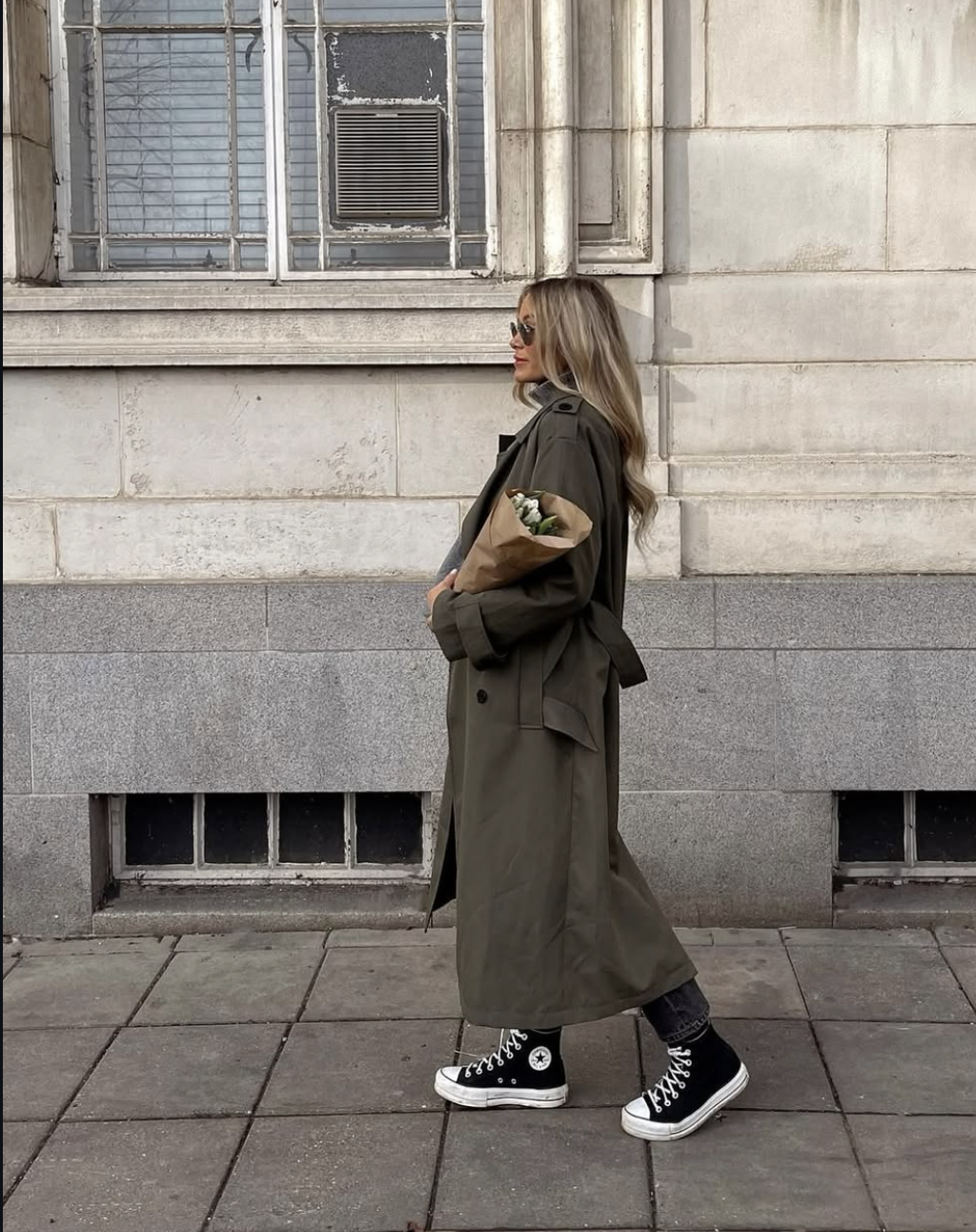 A woman walking in an olive green maxi trench coat styled with black high-top Converse sneakers and carrying a bouquet of flowers in brown paper.