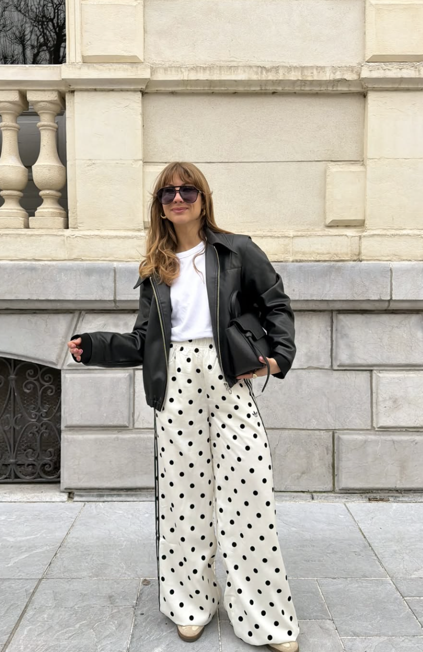 A woman with blonde hair and sunglasses wearing a black oversized leather jacket over a white t-shirt, paired with high-waisted white baggy pants featuring large black polka dots.
