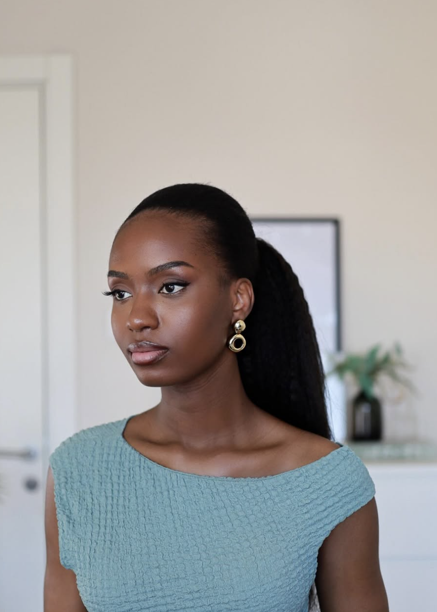 Close-up portrait of a woman showcasing a sleek, low-effort ponytail on dark textured hair, parted cleanly down the middle with smooth edges, wearing a teal knit top and gold hoop earrings.