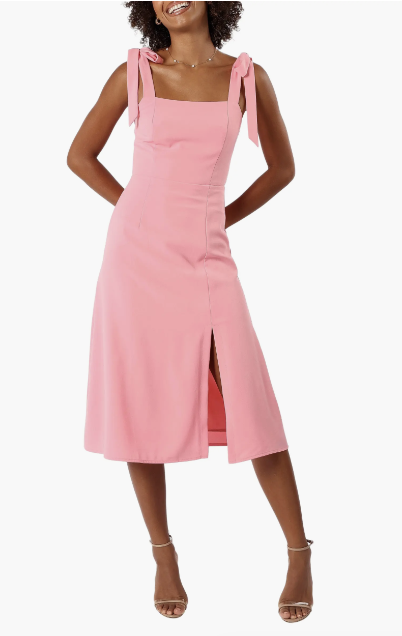 A woman wearing a pink Petal & Pup Laurel A-Line Dress featuring tie-shoulder straps, a square neckline, and a subtle leg slit.