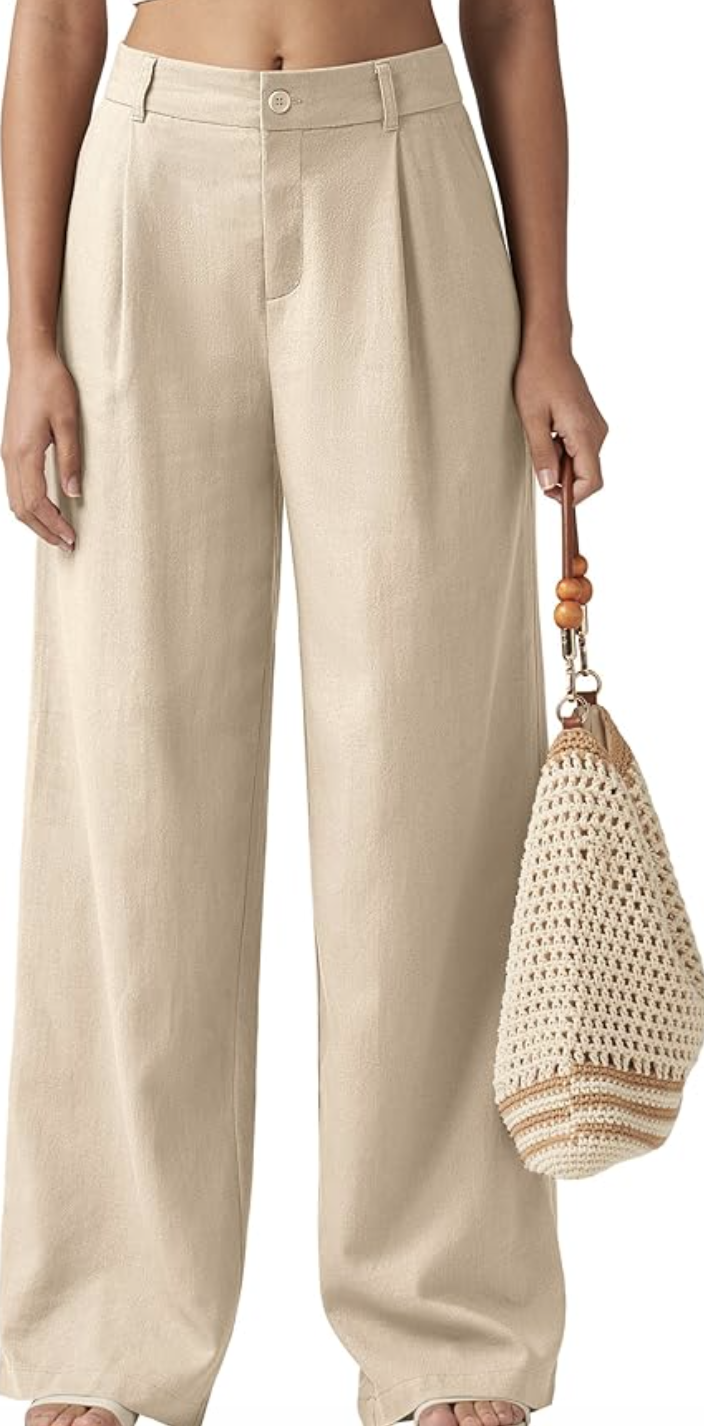 Close-up of a woman wearing high-waisted beige linen wide-leg trousers featuring a tailored waistband, front pleats, and a button closure, holding a straw woven tote bag at her side.