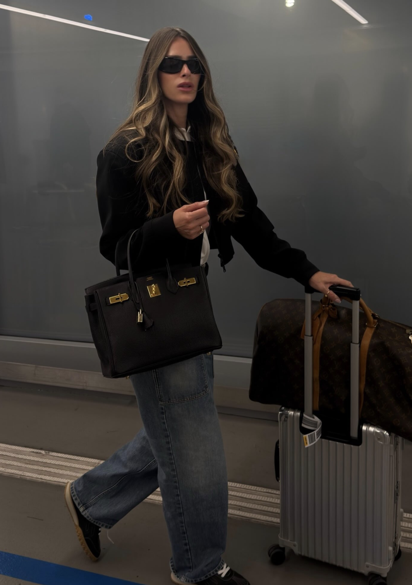 Black bomber jacket with relaxed blue jeans, dark low shoes, a black handbag and wheeled luggage.