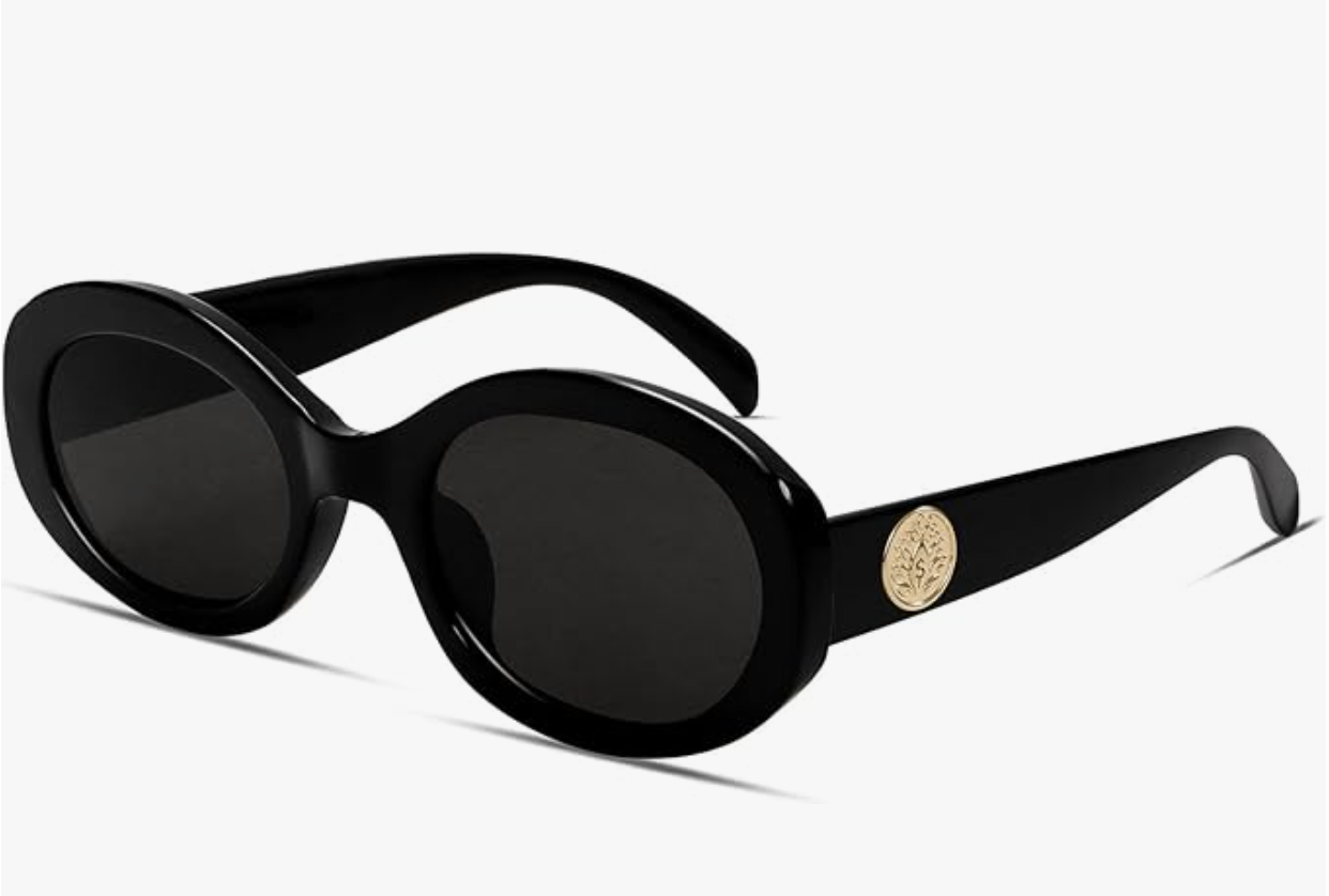 A pair of thick-framed black retro oval sunglasses with a gold designer-inspired emblem on the temple, styled for a luxury vacation look.