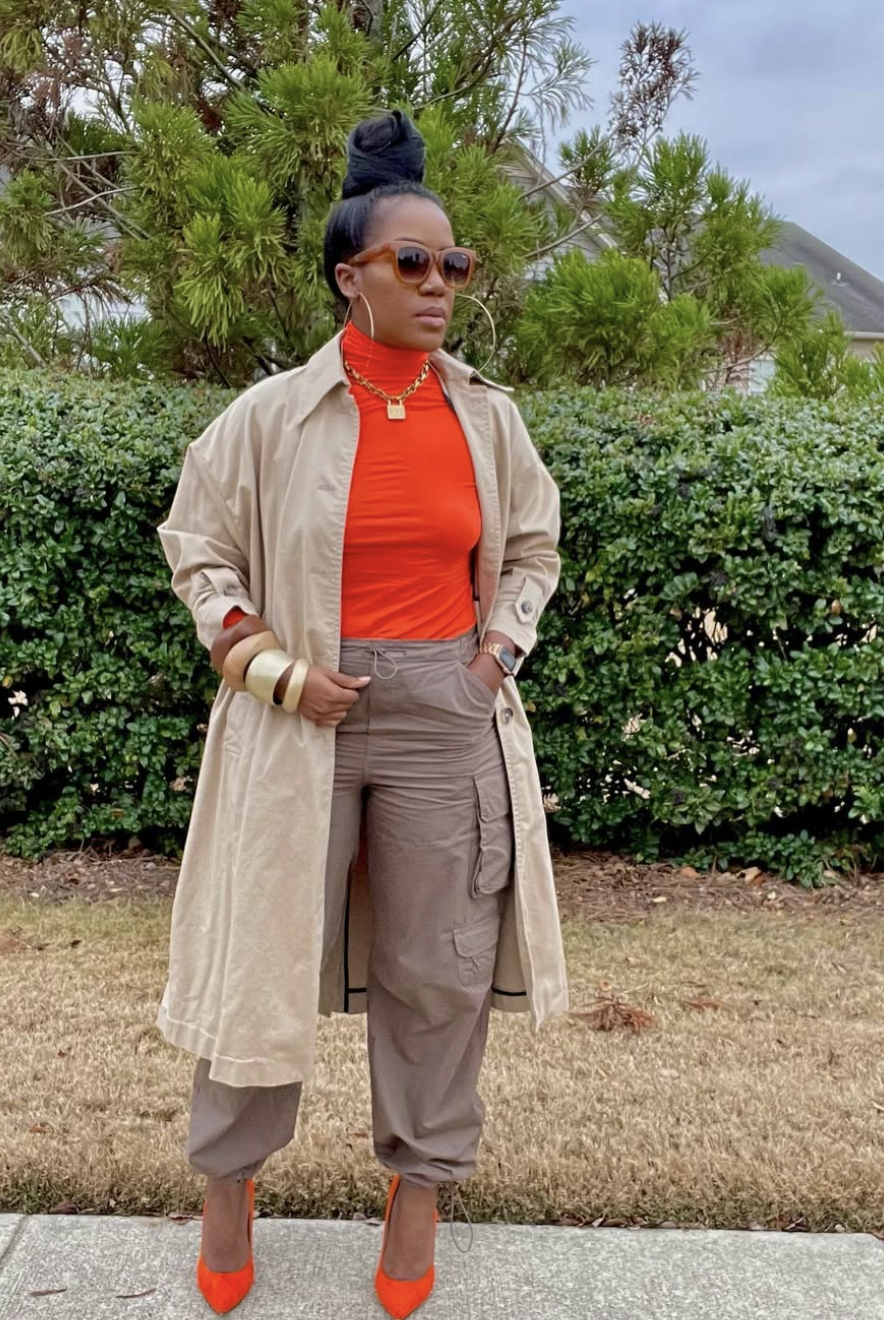 A woman wearing a bright orange turtleneck sweater layered under a tan trench coat, paired with brown trousers and a gold chain necklace.
