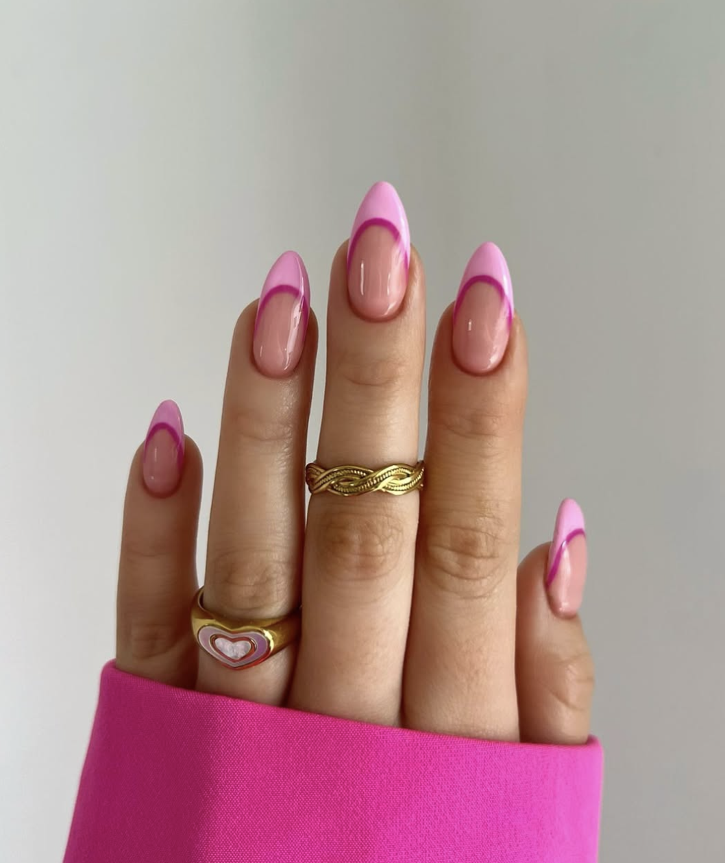 Close-up of a hand showcasing a viral summer manicure with long almond-shaped nails featuring a two-toned neon pink and bubblegum pink French tip design, styled with gold rings and a bright pink sleeve.