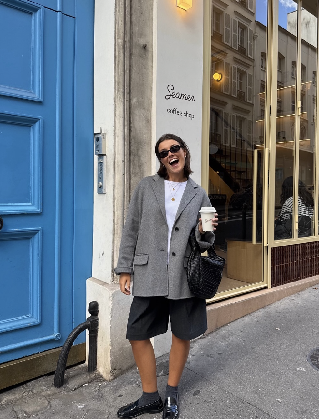 A woman wearing a gray oversized blazer, a white t-shirt, and black tailored Bermuda shorts paired with chunky black loafers and black socks.