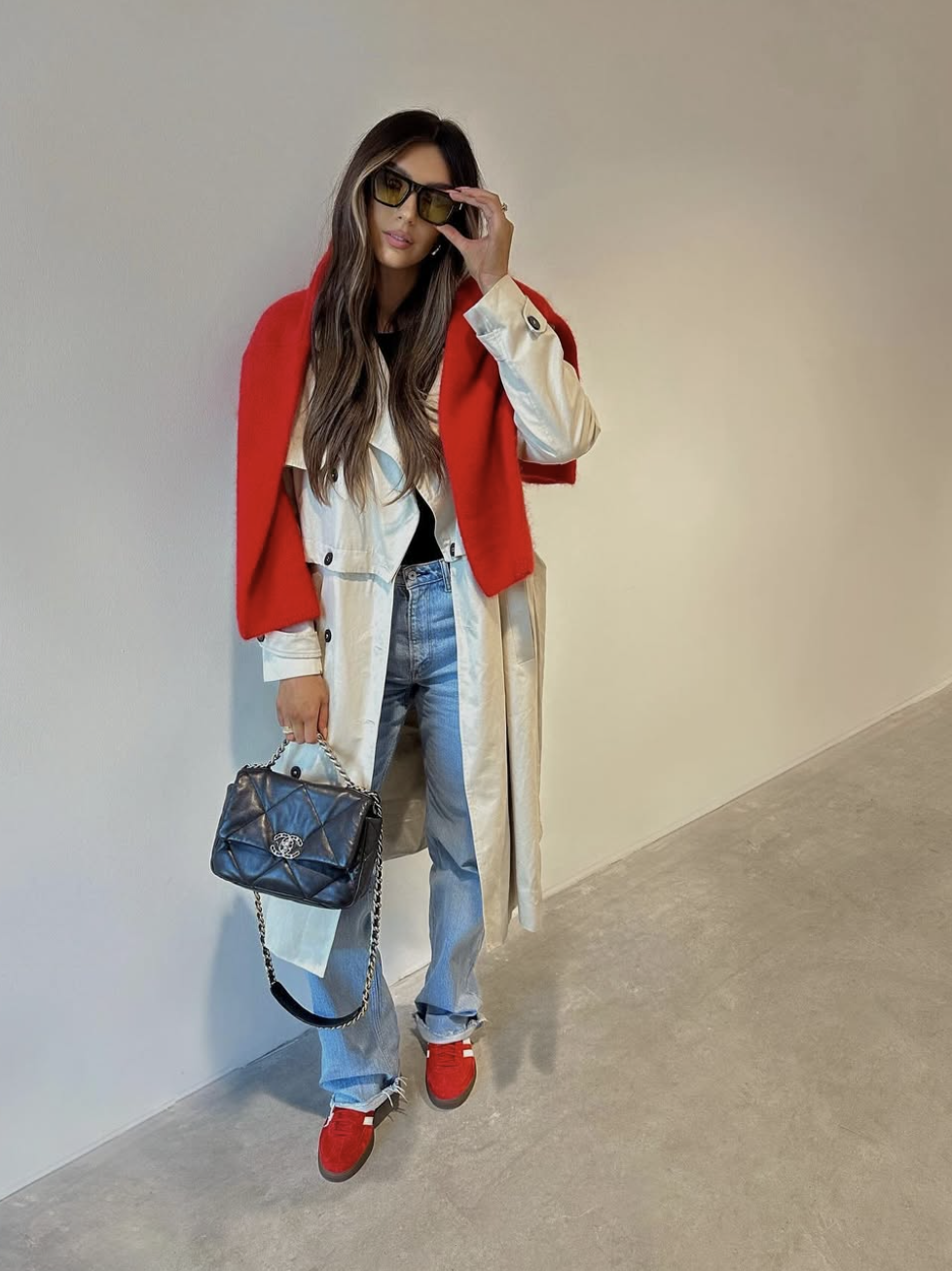 A woman wearing a long white coat over light-wash jeans, styled with bright red sneakers, a red scarf, and a black quilted handbag.