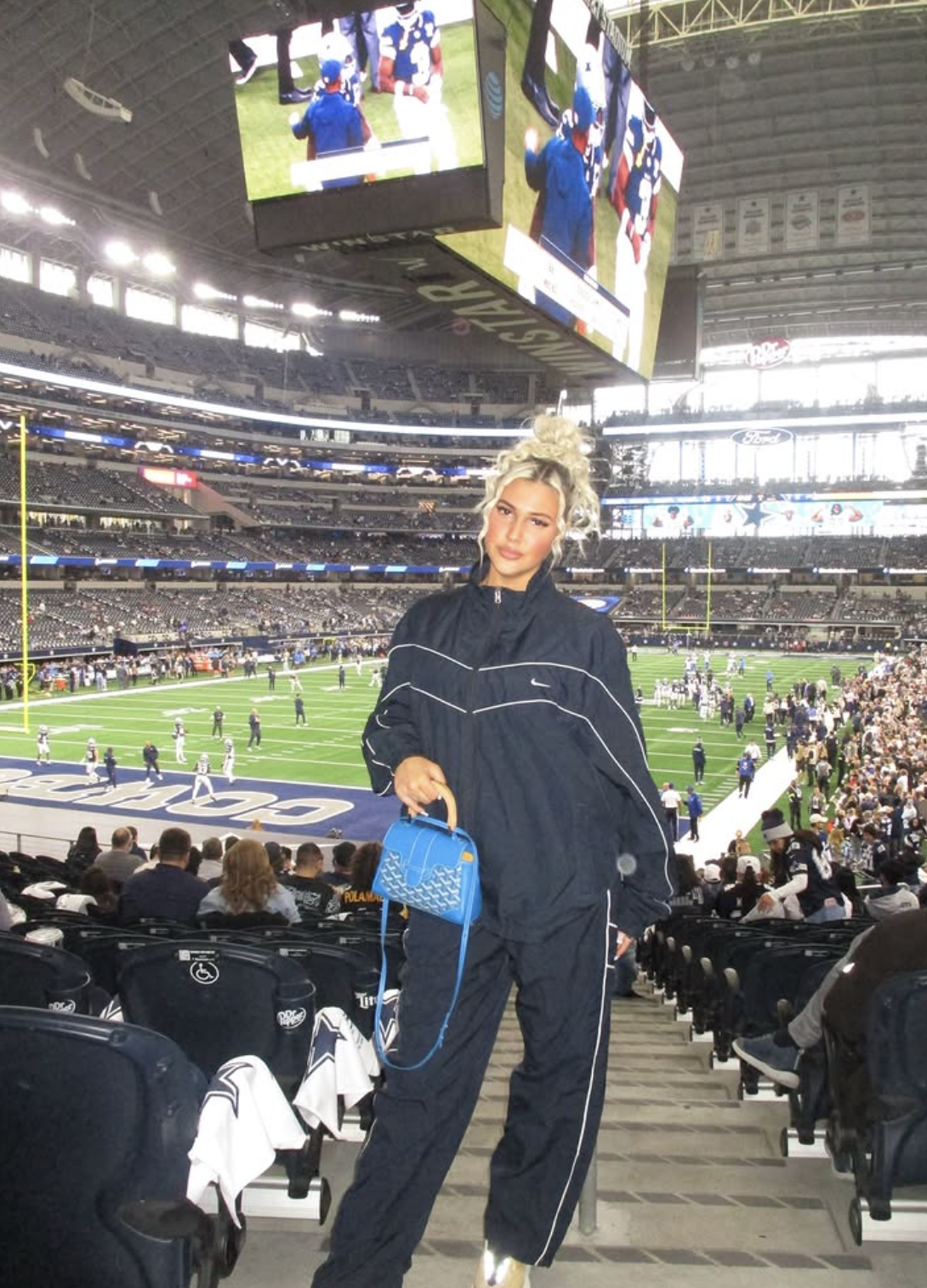 Woman at a football stadium wearing a navy track jacket and matching pants with tan sneakers and a bright blue handbag.