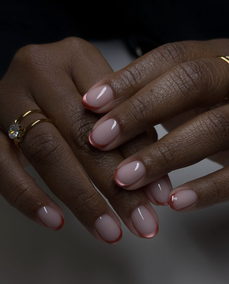 Close-up of short rounded nails featuring a burnt rust-orange micro French tip over a glossy sheer nude pink base.