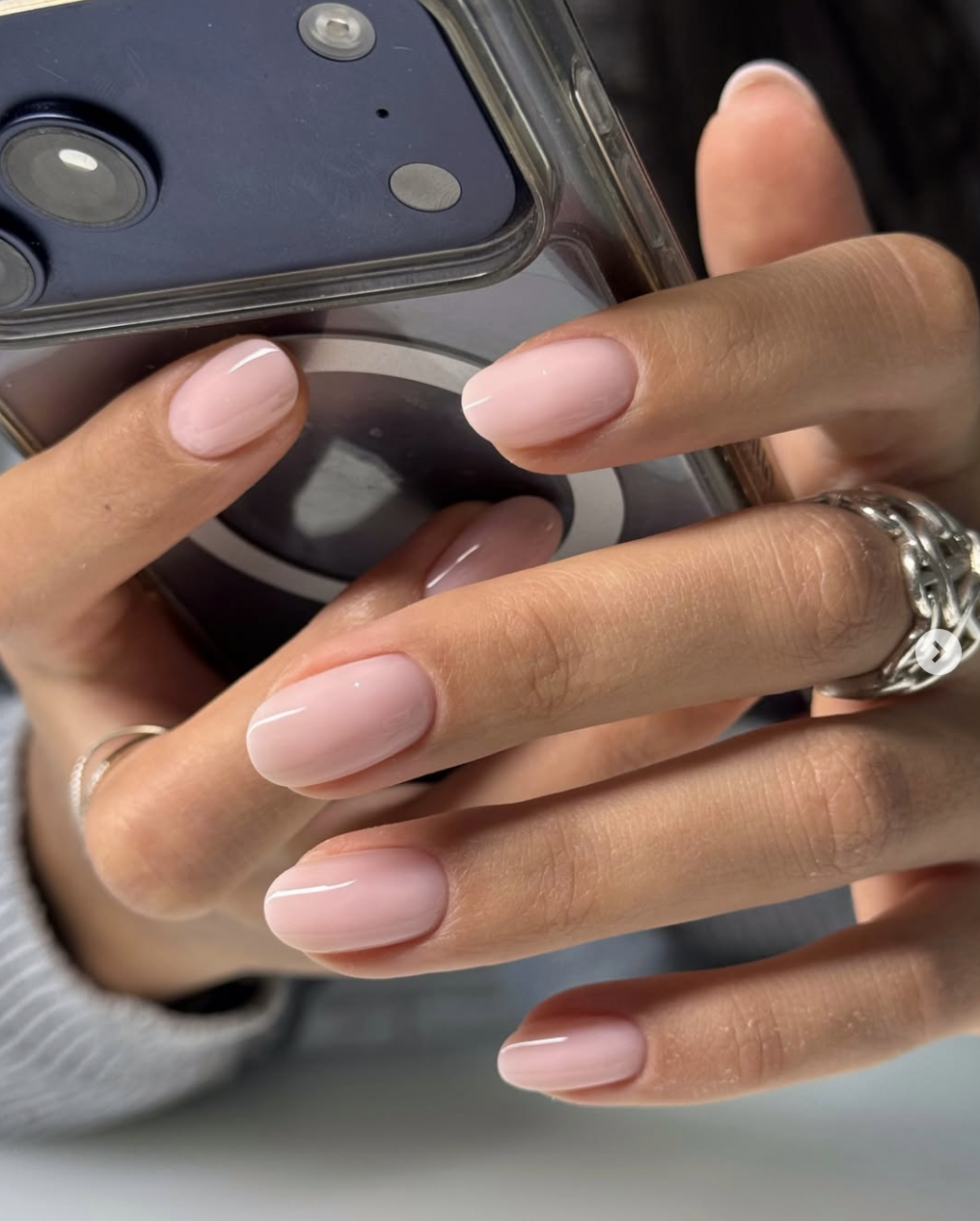 Close-up of a hand showcasing a clean, minimalist summer manicure with short, rounded nails coated in a glossy, sheer pink gel polish while holding a black phone case.