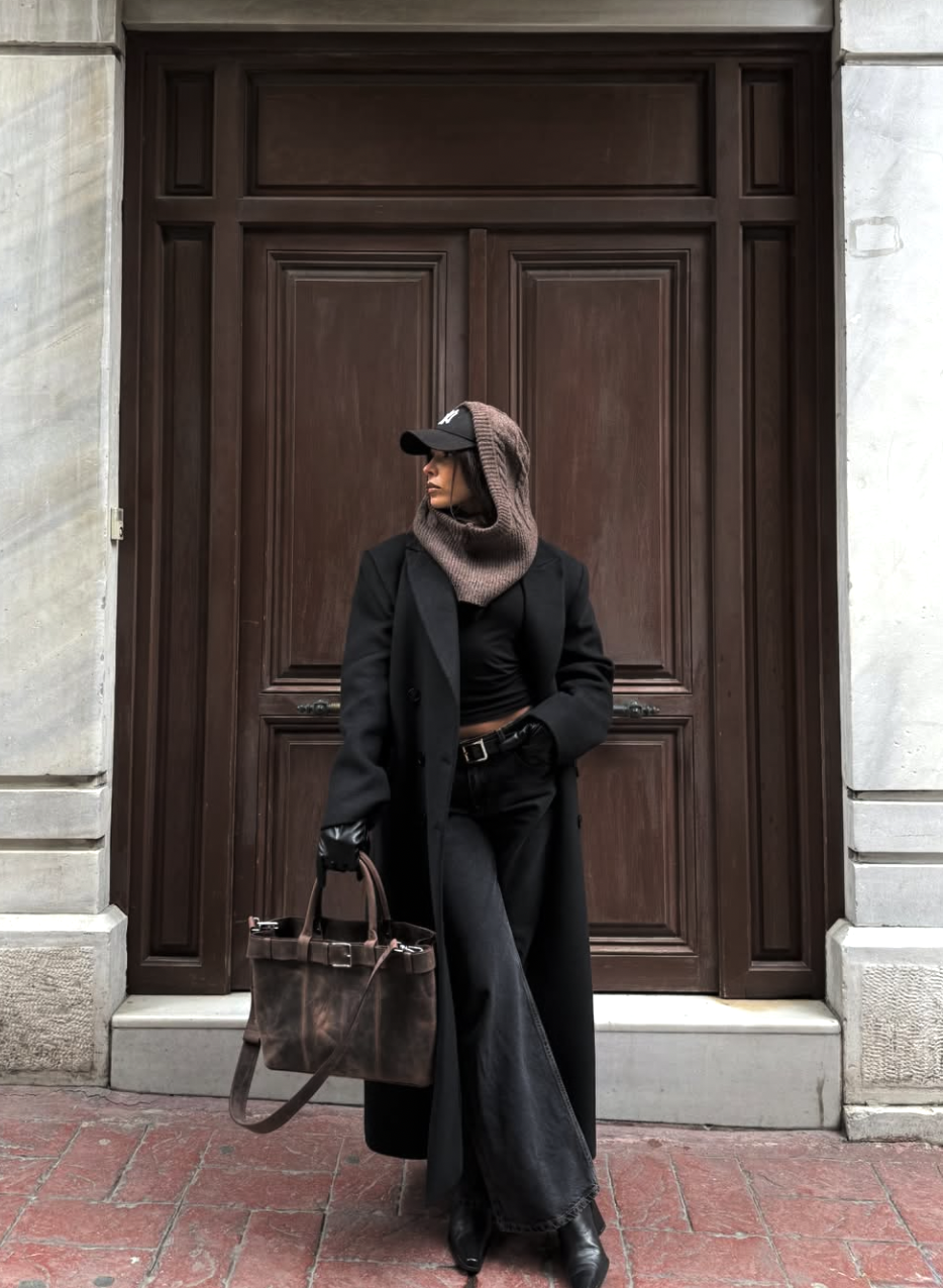 A fashion editor-inspired outfit featuring dark wide-leg jeans paired with sleek black pointed-toe boots, a long coat, and a textured hood, emphasizing long proportions and an intentional silhouette.