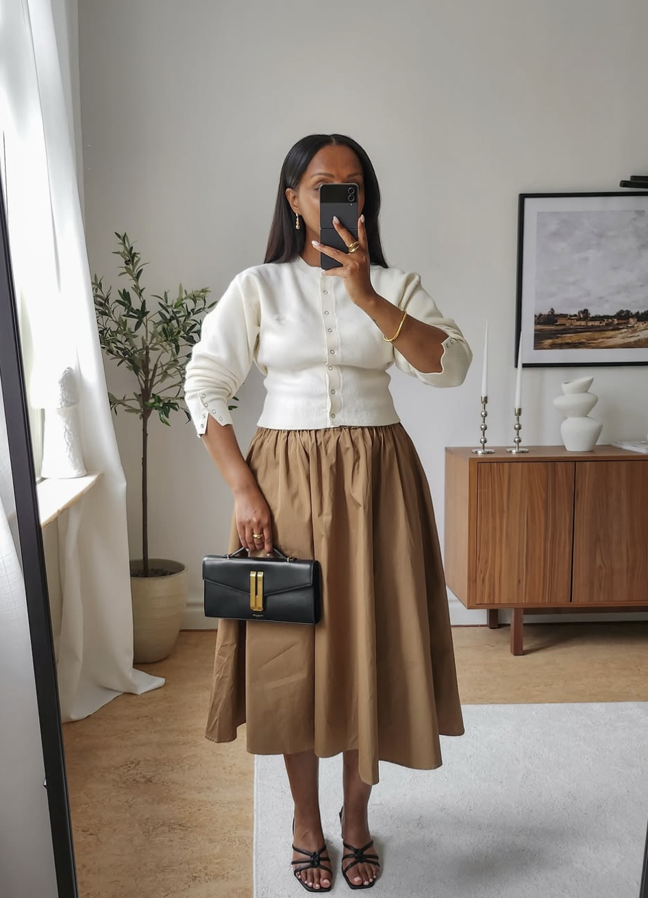 A woman styling a white buttoned-up cardigan as a top with a pleated tan midi skirt and black heels for a professional work outfit.