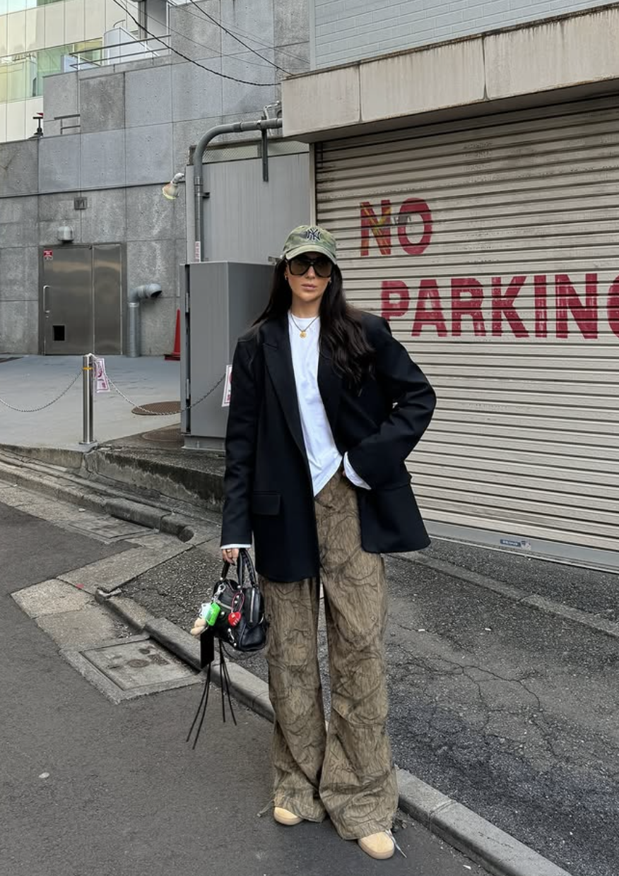 A person wearing a structured black oversized blazer over a white tee, paired with olive green patterned cargo pants, a camouflage baseball cap, and pointed-toe heels.