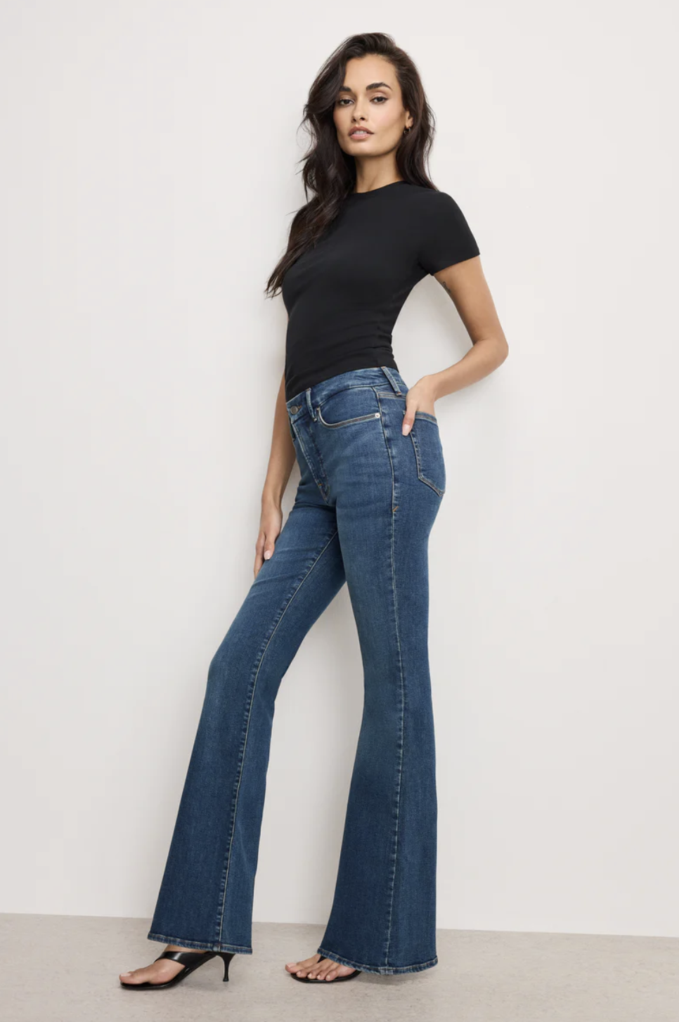 Model wearing Good American Good Legs Flare Jeans in a dark wash, featuring a gap-proof high waistband and a dramatic flare at the ankle to create an hourglass silhouette for an athletic figure.