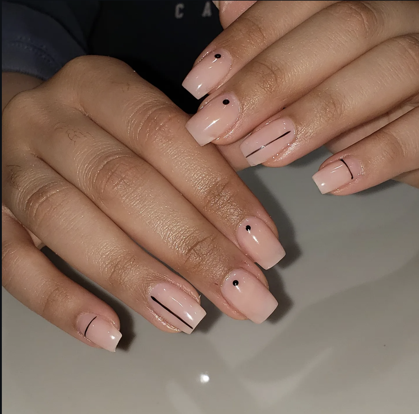 Short square nails with a glossy pale pink base, featuring minimal black dots on the center of the cuticles and thin black vertical line accents.