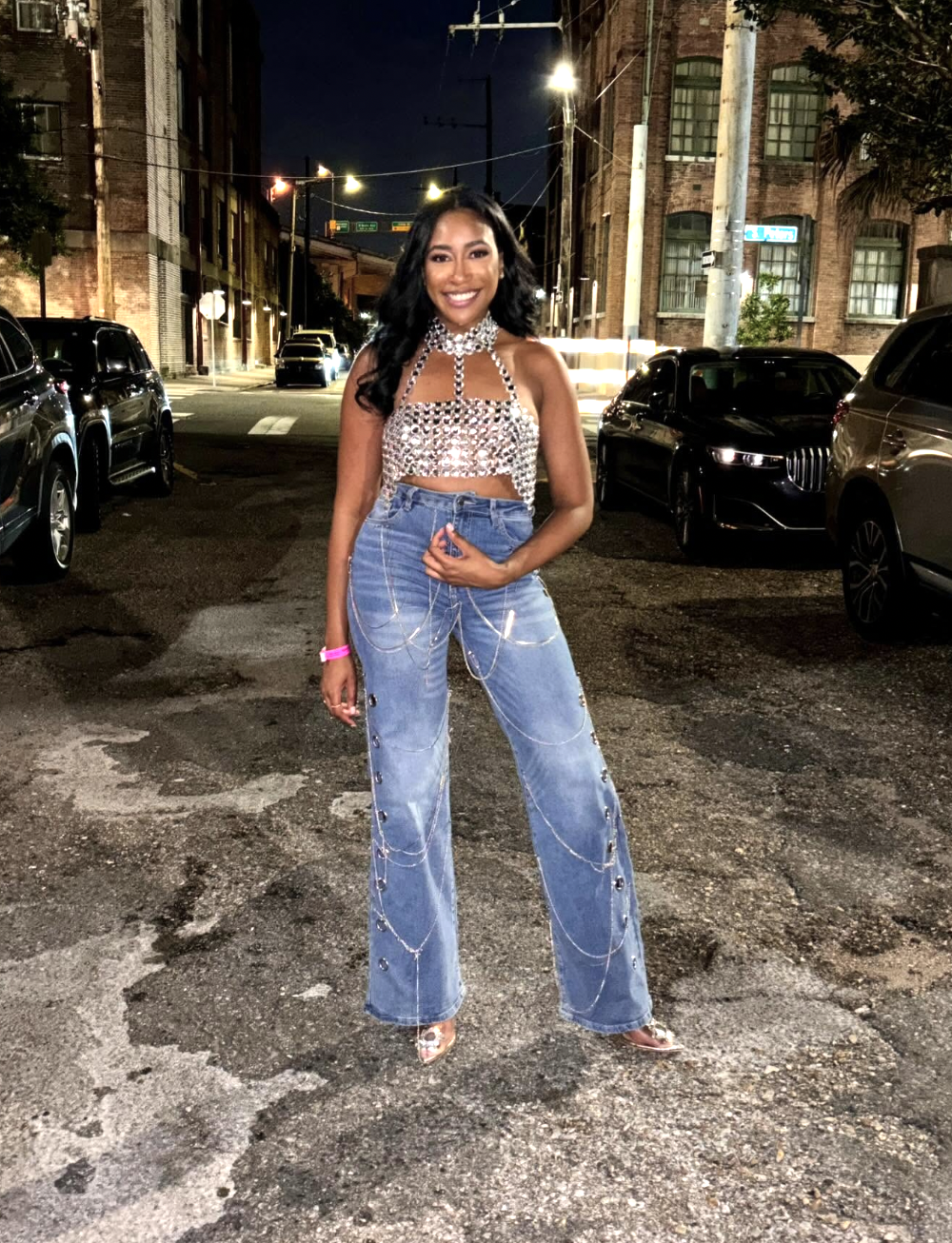 A woman standing outdoors at night wearing a sparkling silver crop top and high-waisted, wide-leg denim jeans decorated with metal grommets and layered silver body chains draped across the legs.
