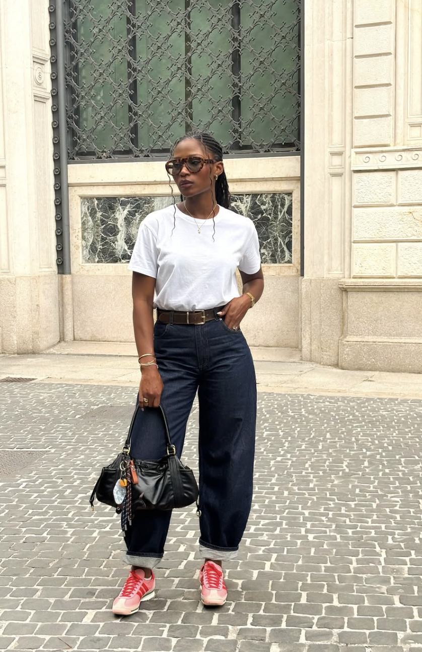 A woman styling dark-wash barrel-leg jeans with a tucked-in heavyweight white crewneck t-shirt, a brown leather belt, and red retro sneakers.