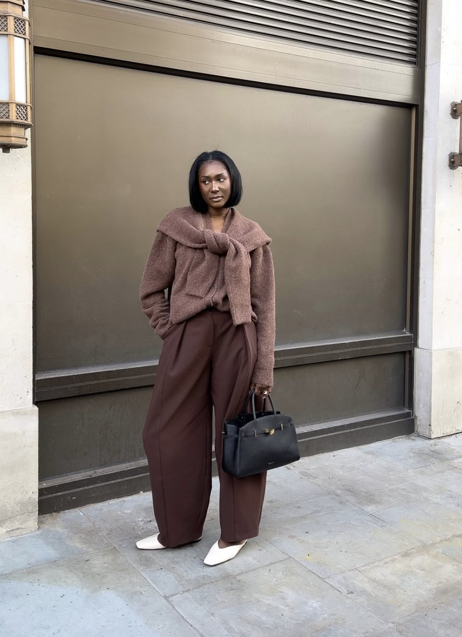 A woman wearing a monochromatic chocolate brown outfit featuring an oversized knit sweater with a matching sweater draped over her shoulders and high-waisted wide-leg trousers, styled with a black leather handbag.