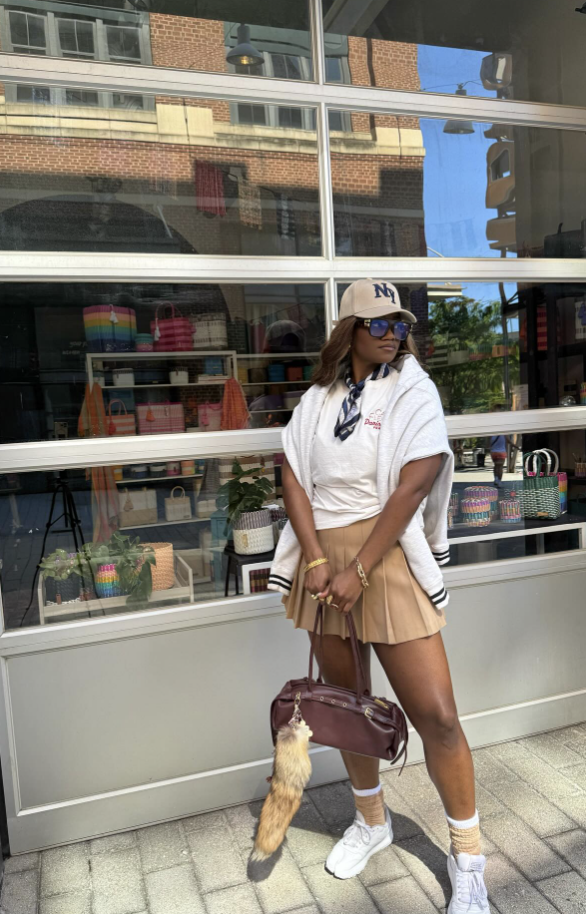 Woman wearing a white oversized shirt, camel pleated mini skirt, baseball cap, crew socks and sneakers.