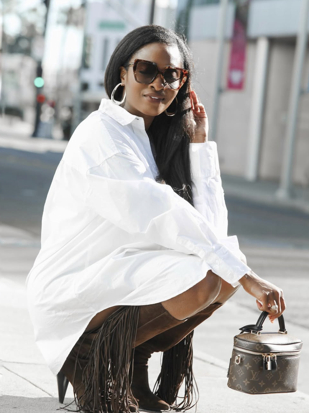 A woman sitting outdoors wearing an oversized white button-down shirt dress and tall brown suede boots with fringe detailing along the sides.