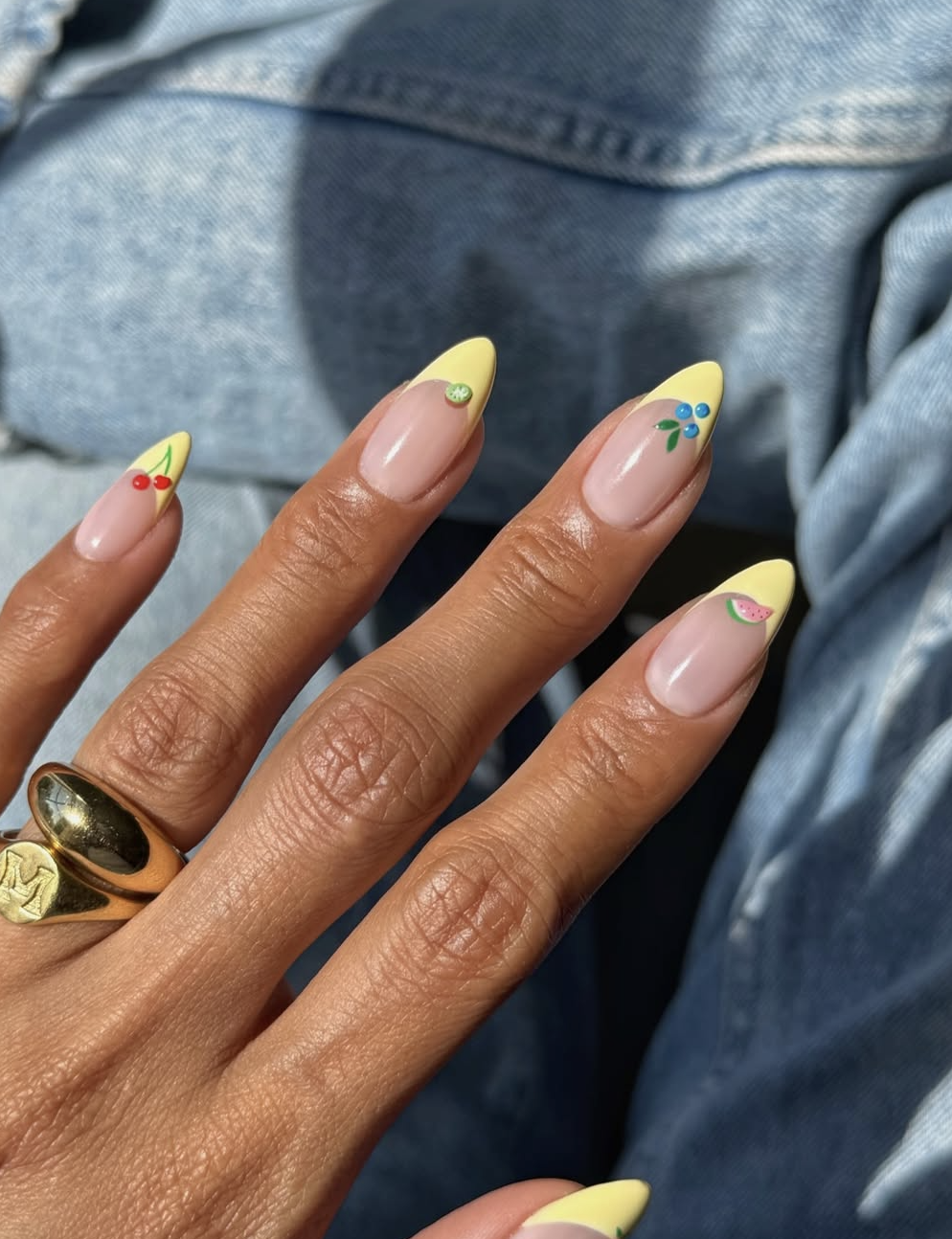 A close-up of almond-shaped nails with a light butter-yellow French manicure accent, featuring tiny 3D cherry, lime, blueberry, and watermelon nail art on a clear base, resting against denim.