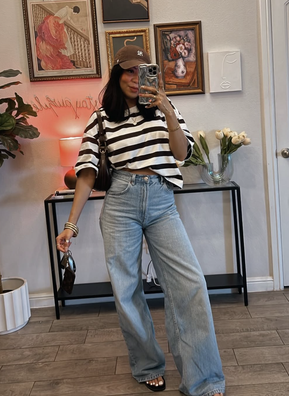 A woman taking a mirror selfie wearing an oversized black and white striped t-shirt tucked into high-waisted, light-wash wide-leg denim jeans, styled with a brown New York Yankees baseball cap and a dark shoulder bag.