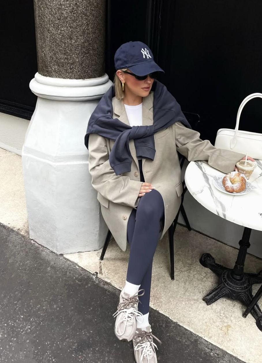 Woman wearing dark stirrup leggings with an oversized beige blazer, a navy sweatshirt tied over her shoulders, a New York Yankees baseball cap, and chunky white sneakers.