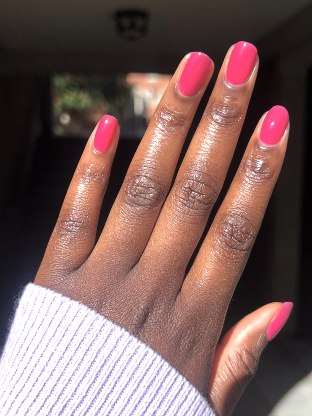 A close-up of long, square-shaped nails with a high-shine, translucent hot pink jelly manicure on a person with a deep skin tone, wearing a lavender knit sleeve.