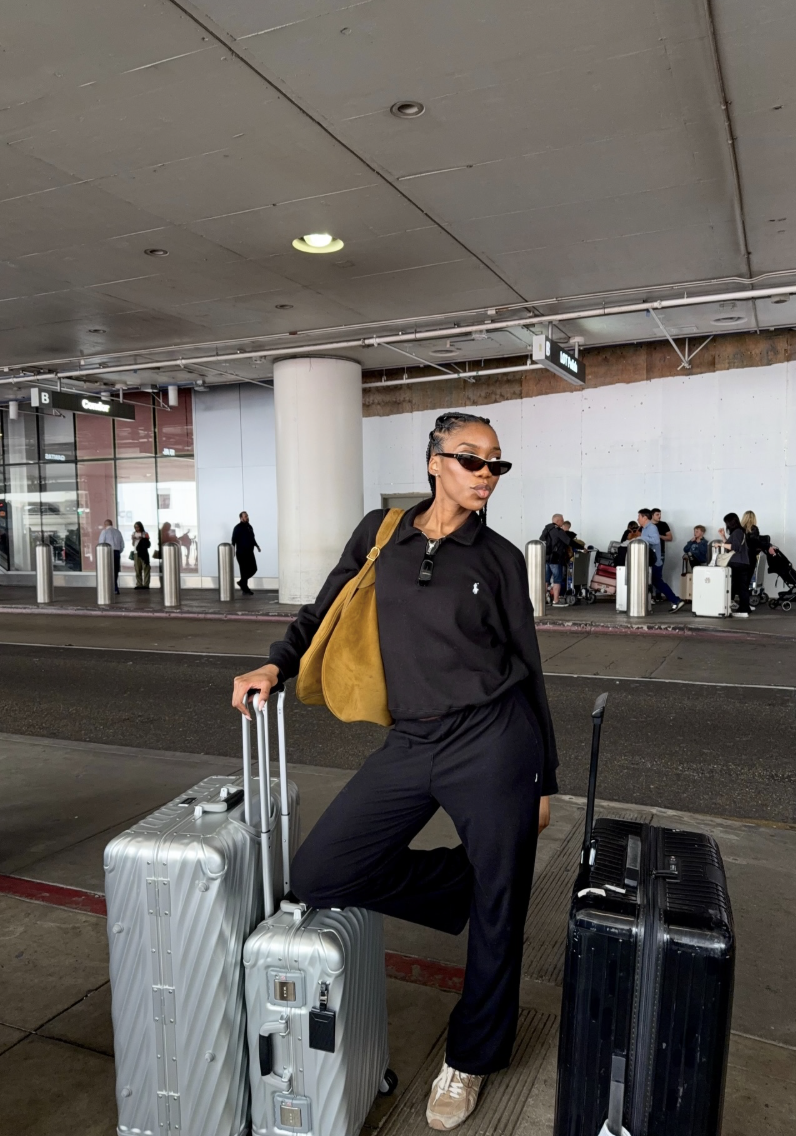 Dark coordinated tracksuit with light shoes and a mustard shoulder bag beside suitcases.