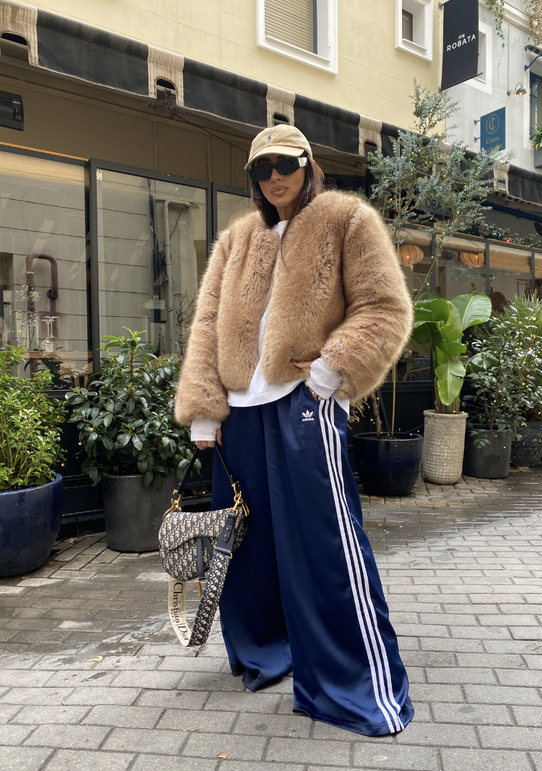 Professional style hack: Navy Adidas Firebird loose track pants styled with a cropped tan faux fur coat, a beige baseball cap, and a Dior saddle bag for a luxurious Sportif Luxe spring outfit.