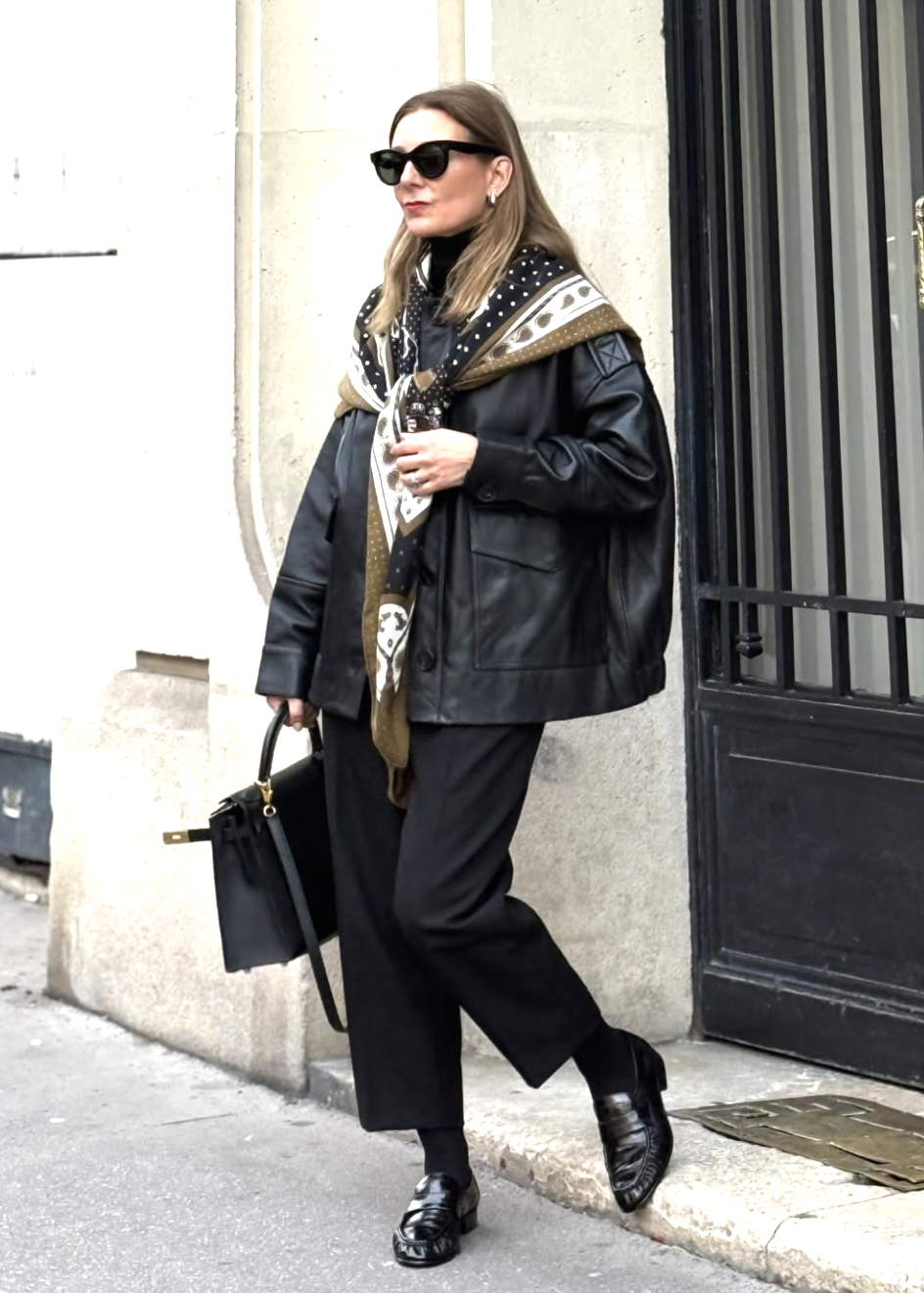 A woman wearing an all-black minimalist outfit featuring a masculine leather jacket layered over a black turtleneck and trousers with a black cashmere shawl and polished loafers.