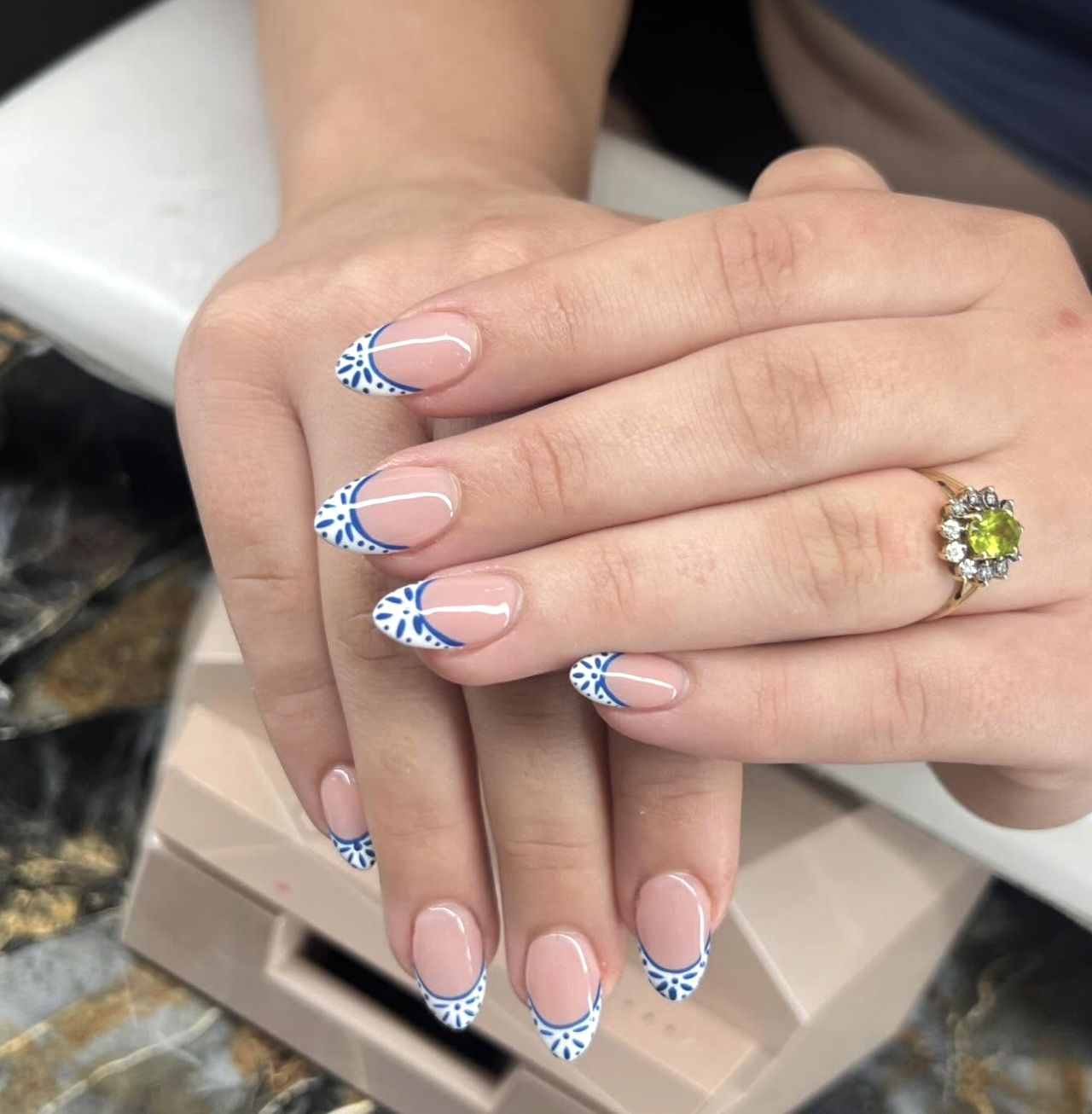 Long almond-shaped nails with a sheer nude base and cobalt blue French tips featuring white intricate floral and tile patterns inspired by Mediterranean porcelain.