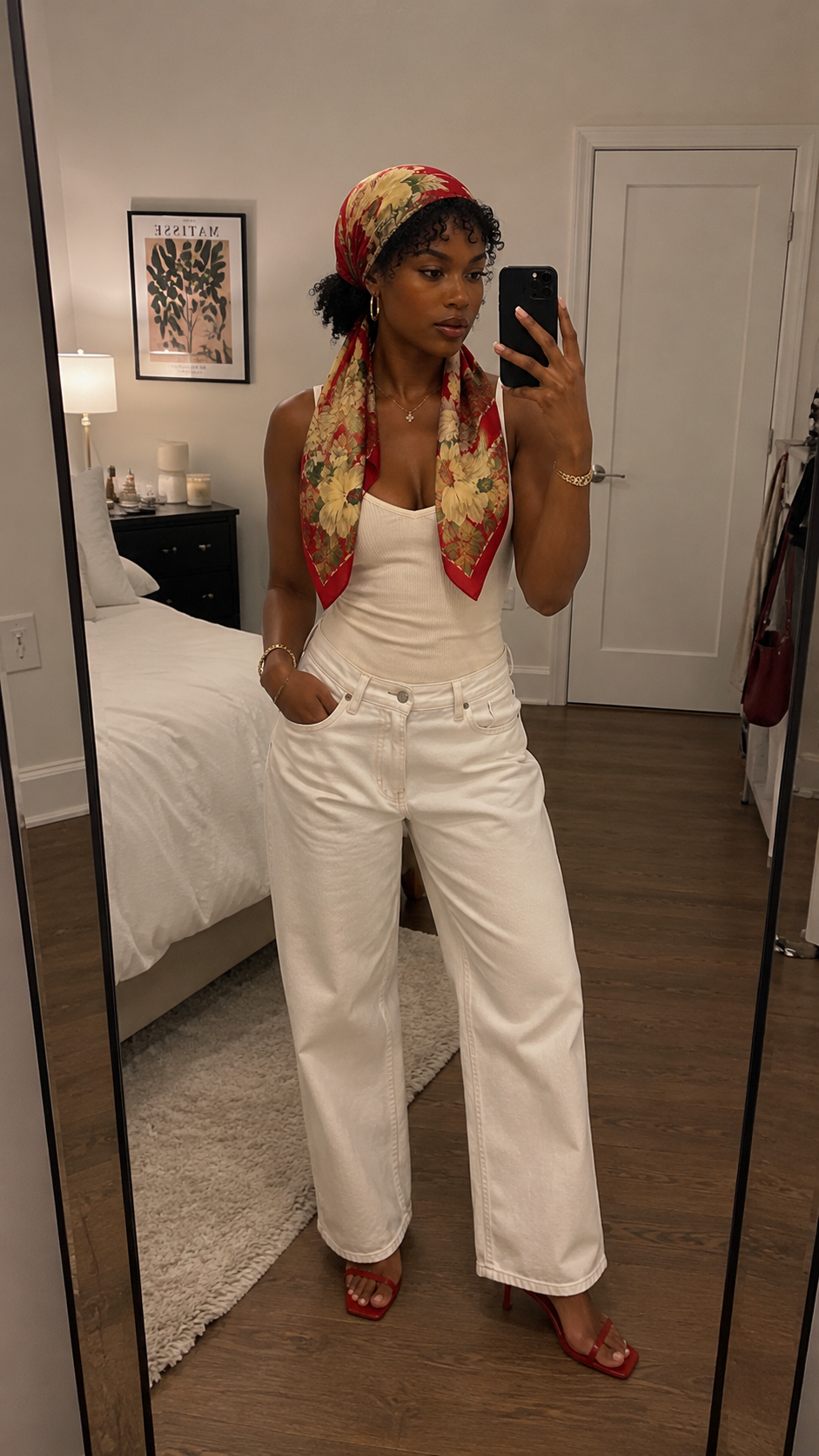 A woman wearing a red floral vintage silk headscarf tied bandana-style over dark curly hair, paired with a white top and cream pants.