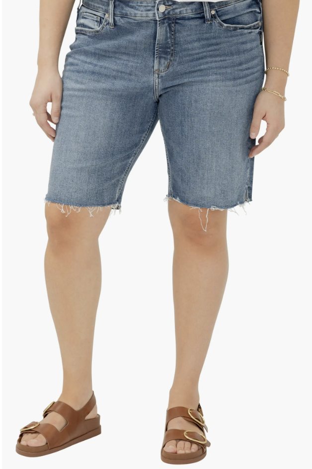 A woman wearing dark-wash Silver Jeans Co. Elyse curvy denim Bermuda shorts with a raw hem, designed to fit thick thighs and an apron belly without a waist gap.
