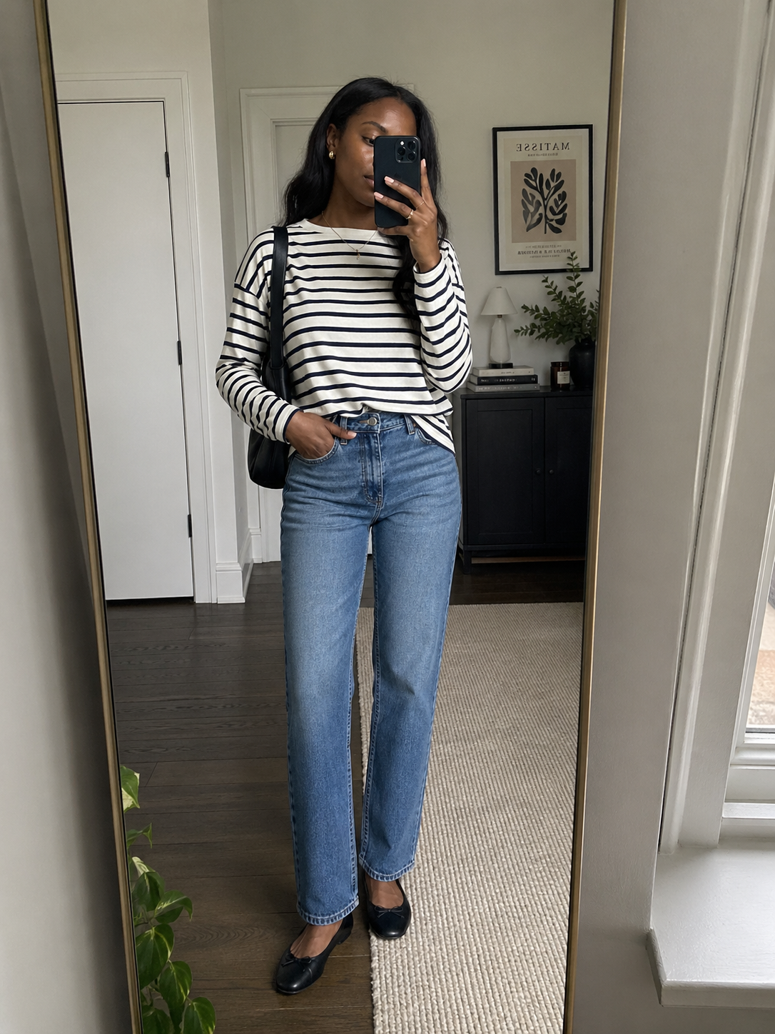: A woman taking a mirror selfie wearing a classic black and white striped Breton tee tucked into high-waisted straight-leg jeans.