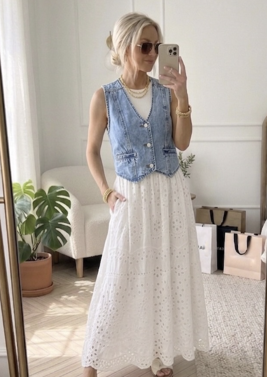 A woman styling a casual summer outfit featuring a light-wash denim vest layered over a white eyelet midi dress, paired with gold layered necklaces, a gold bracelet, and aviator sunglasses.