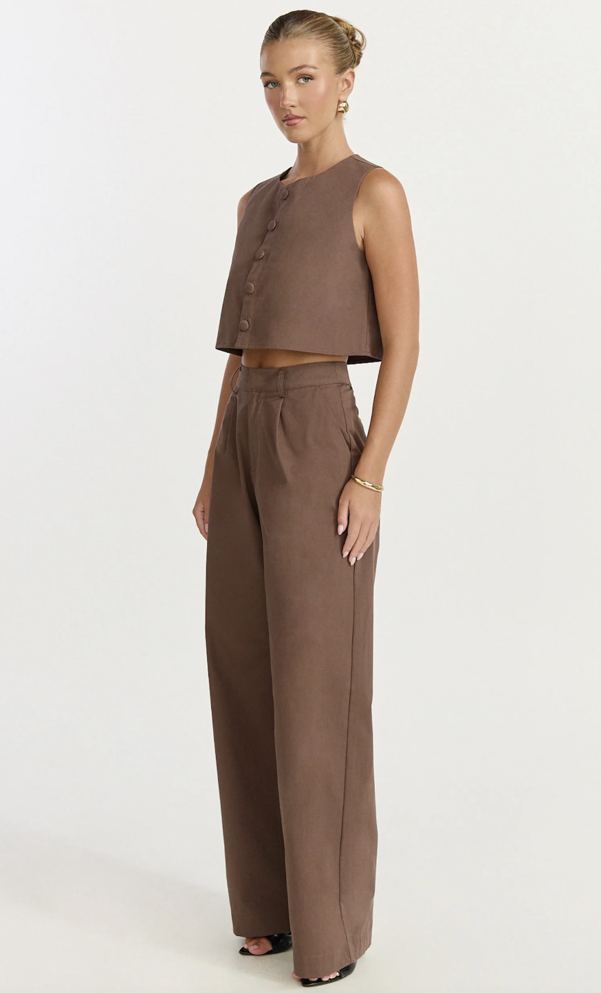 A product view of a woman wearing a monochromatic chocolate brown linen outfit featuring a cropped button-up vest top and high-waisted pleated wide-leg trousers.