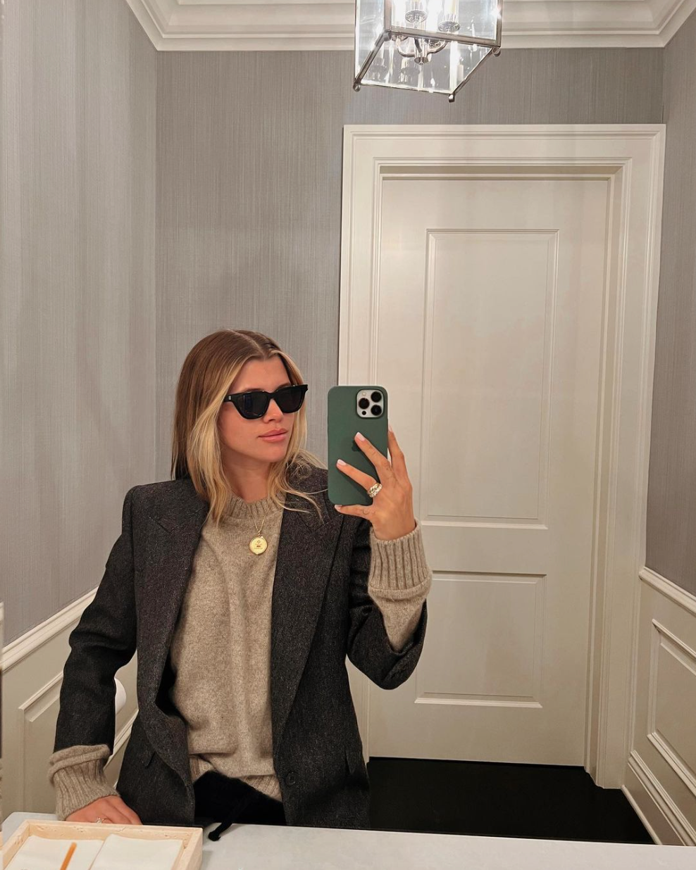 Sofia Richie taking a mirror selfie wearing a structured charcoal blazer over a neutral cashmere sweater and gold pendant necklace, embodying the Quiet Money aesthetic.