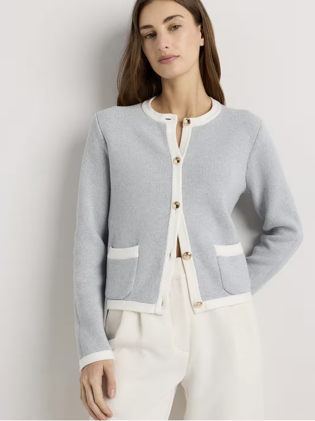 A woman wearing a light blue structured knit cardigan jacket with gold crest buttons paired with high-waisted cream trousers.