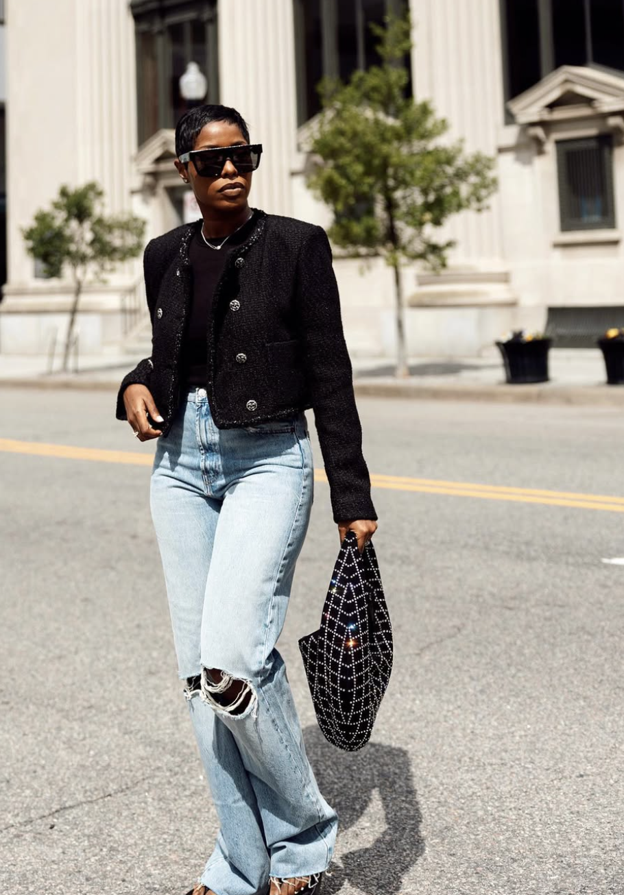 A woman wearing light blue distressed jeans with a black cropped tweed jacket and strappy brown leather sandals, carrying a black crystal-embellished net bag.