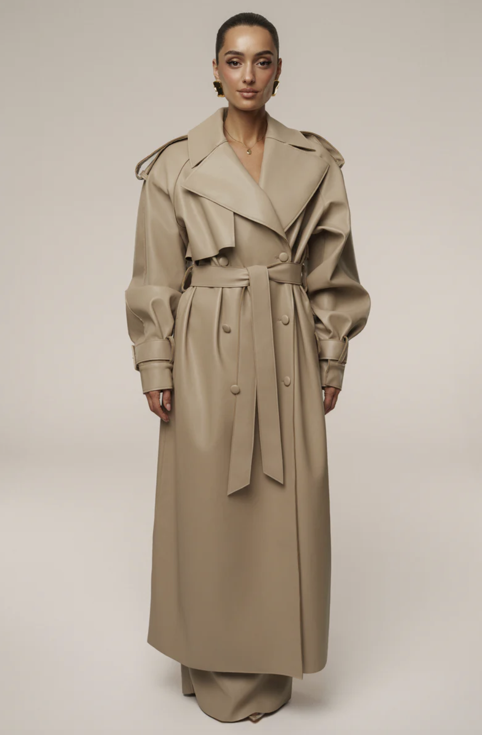 A woman wearing a long tan faux leather trench coat with a cinched waist belt and double-breasted buttons.