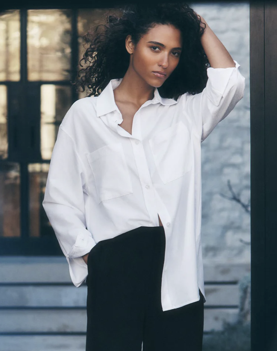 A woman with curly hair wearing an oversized, crisp white Alo Yoga Dreamscape button-down long-sleeve shirt, tucked loosely into dark bottoms.